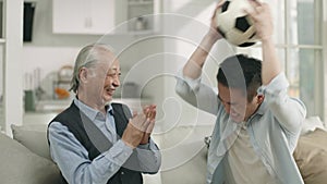 asian senior father and adult son watching football game on tv at home