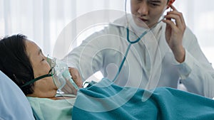 Asian senior female patient with oxygen mask lying on bed in hospital with asian male doctor examining patient with stethoscope