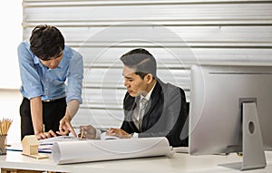 Asian senior good-looking boss in suit sitting at desk and working with junior employee in office with roll of paper. Businessmen