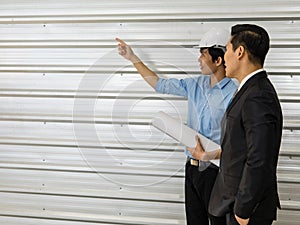 Asian senior good-looking boss in suit standing and working with junior employee wearing white safety helmet and holding roll of