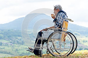 Asian senior man drink tea from a cup and sit on wheelchair with near the cliff and mountain and grassfield as background