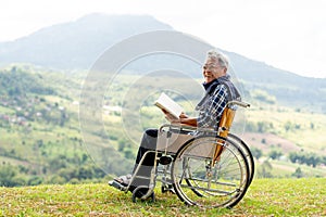 Asian senior man with eyeglasses hold the book and sit on wheelchair also look at camera and stay near the cliff with grassfield