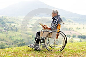 Asian senior man with eyeglasses read the book and sit on wheelchair also stay near the cliff with grassfield and mountain as