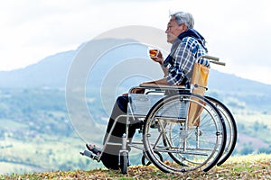 Asian senior man hold a cup of tea and sit on wheelchair with lood forward and stay near the cliff and mountain and grassfield as