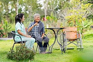 Asian senior man hold a glass of water and talk to nurse after take the medicine from her and they sit outdoor in the garden with