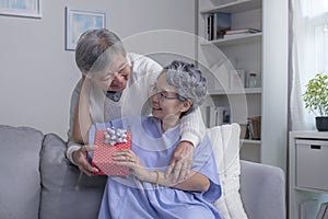 Asian senior man husband giving present gift box for Valentine`s day to happy surprised wife, sitting together on couch at home