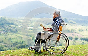 Asian senior man sit on wheelchair and read book also stay near cliff with meadow and mountain in the background with warm light
