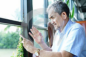 Asian senior man sitting among nature atmosphere, talking and waving to someone by online video call on mobile smartphone