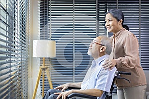 Asian senior man sitting in wheelchair at home looking out the window with his wife is supporting and caring for love and