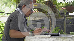 Asian senior man using laptop and writing on notepad at home.