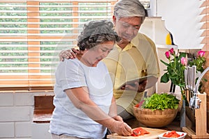 Asian senior wife slicing tomatoes on wooden cutting  board and using tablet computer to searching menu recipe.senior with