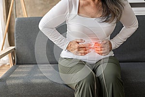 Asian senior woman having stomach pain while sitting on sofa at home,
