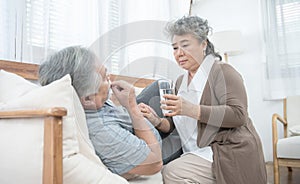 Asian Senior woman look after give glass of water to elder man taking medicines and drinking water while lie on couch at home,