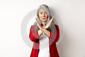 Asian senior woman in red blazer making cross gesture, pleading you stop, disagree and disapprove something, standing