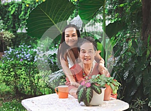 Asian senior woman sitting with her daughter  at white table in beautiful garden, daughter hugging her mother from the back,