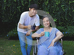 Asian senior woman sitting on wheelchair, drinking coffee or tea with her son in the garden. looking at camera, smiling happily