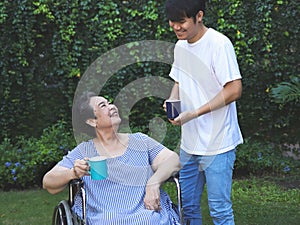 Asian senior woman sitting on wheelchair, drinking coffee or tea with her son in the garden. smiling happily