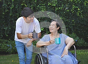 Asian senior woman sitting on wheelchair, drinking coffee or tea with her son in the garden. smiling happily