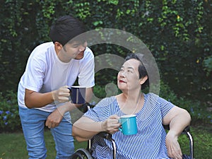 Asian senior woman sitting on wheelchair, drinking coffee or tea with her son in the garden. smiling happily