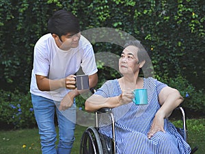 Asian senior woman sitting on wheelchair, drinking coffee or tea with her son in the garden. smiling happily