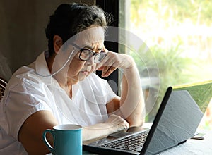 Asian senior woman sitting by the windor with blue cup of coffee and computer laptop on table , looking thoughfully at computer
