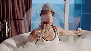 Asian senior woman takes, drinks cocktail in warm bubble bath. Waiter brings a drink to rich elderly female in foam