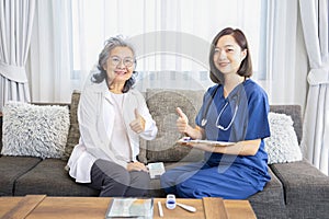 asian senior woman and young female nurse sitting on couch and doing thumb up while a nurse visiting patient at home