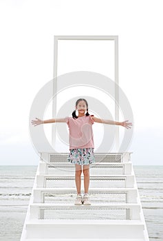 Asian smiling girl child arms wide open standing on open door gate with stairs to the sea and sky. Kid hands outstretched or