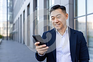 Asian smiling young man walking down the street near an office building and using a mobile phone