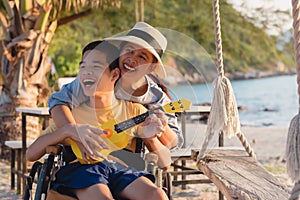 Asian special child on wheelchair is singing, playing ukulele happily on the beach
