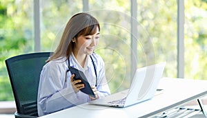 Asian successful professional young female doctor in white lab coat uniform with stethoscope sitting smiling using typing laptop