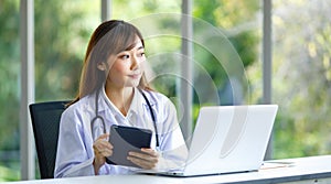 Asian successful professional young female doctor in white lab coat uniform with stethoscope sitting smiling using typing laptop