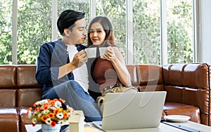 Asian sweet couple boyfriend and girlfriend sitting on sofa in cozy living room at home in morning, drinking coffee together for