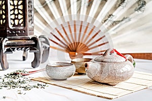 Asian teapot with teacups on bamboo tablamat decorated with chinese fan, lantern and scattered green tea on white marble
