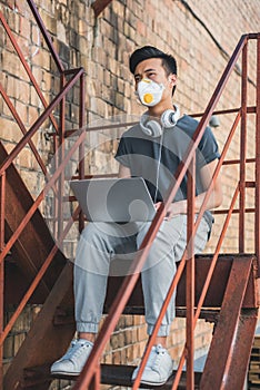 asian teen in protective mask sitting with laptop on staircase and looking away air