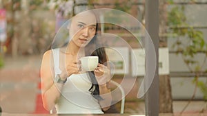 Asian teenage girl holds a coffee cup in a cafÃ© and looks at the outside camera
