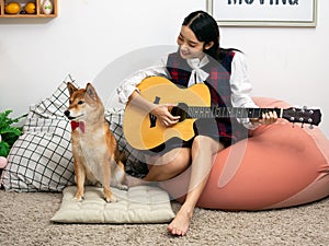 Asian teenage playing guitar with Shiba Inu dog sitting in white living room