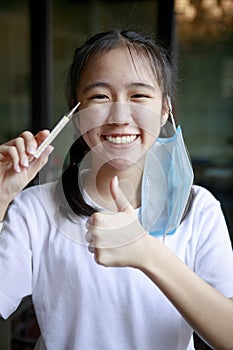 Asian teenager toothy smile with protection mask hanging beside head and themometer in hand
