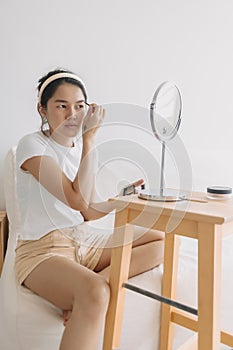 Asian Thai woman using pencil and drawing eyebrow, sitting on white couch in front of the mirror in room apartment