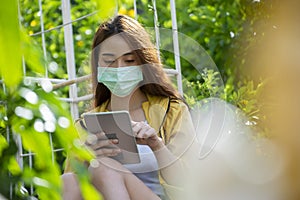 Asian Thai Woman wear mask using tablet and touch screen via smartphone in business market at park. Working online from Home and