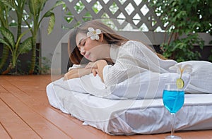 Asian Thai young woman lying and relaxing on bed near swimming pool in spa salon with a drink glass, beauty and body care concept