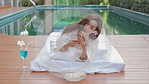 Asian Thai young woman lying and relaxing, using mobile phone on bed near swimming pool in spa salon with a drink glass, beauty