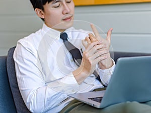 Asian tired overwork male businessman employee in business outfit sitting on sofa working with laptop computer holding hand
