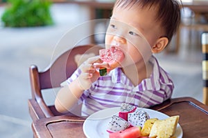 Asian toddler boy sitting in high chair using hands eating watermelon, pineapple, dragonn fruit