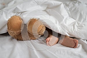 Asian toddler feet beside teddy bear feet in white bed, sheet and pillow