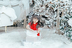 Asian toddler girl in red dress opening gift boxes with presents under Christmas tree at home