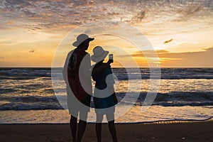 Asian tourist couple taking pictures of sunset at the beach, Beautiful sunset above sea, Colorful Orange and golden sunset sky,