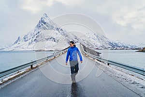 An Asian tourist man walking on road and traveling in Lofoten islands, Norway, Europe. White snowy mountain hills, nature