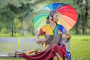 Asian transgender university student holding rainbow pride umbrella sitting on bench at campus park