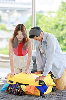 Asian traveler couple in casual vacation outfit with sunglasses and hat sitting smiling on carpet floor in living room packing